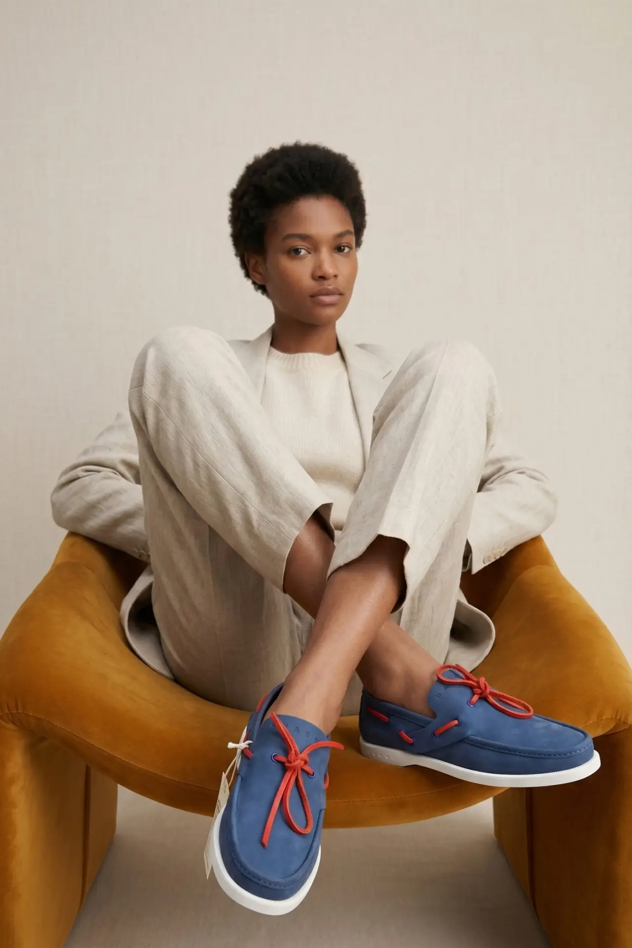 Professional editorial shot of a model wearing premium navy blue sneakers and a cream linen suit, sitting in a mustard velvet chair. Part of the Shoeaholic luxury footwear collection in Bahrain. || صورة عصرية لعارض أزياء يرتدي أحذية رياضية كحلية فاخرة مع بدلة كتان، من تشكيلة متجر شوهوليك للأحذية الراقية في البحرين والخليج.