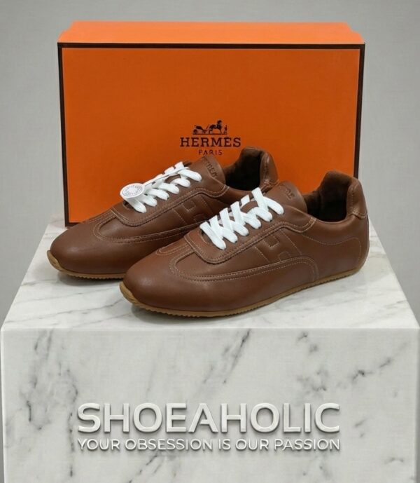 حذاء رياضي من الجلد البني الفاخر مع صندوق العلامة التجارية، يعكس الأناقة والجودة العالية في البحرين. ||| Premium brown leather sneakers displayed with a signature luxury brand box on a minimalist background.